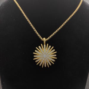 May include: A gold necklace with a sunburst pendant. The pendant has a central cluster of small, clear stones surrounded by radiating gold spikes. The chain is a box-style design. The necklace is displayed against a black background.