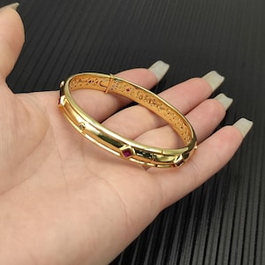 Op de afbeelding: Een goudkleurige armband met een sluiting, met kleine rode en heldere edelstenen. De armband wordt in een hand gehouden, waarbij het gepolijste oppervlak en het ingewikkelde ontwerp worden getoond. De armband heeft een herhalend patroon aan de binnenkant.