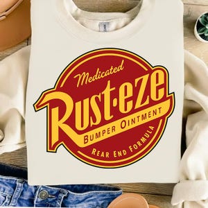 May include: Cream-colored sweatshirt featuring a large, circular graphic with the text "Medicated RUST-EZE Bumper Ointment Rear End Formula" in red and yellow. The design is reminiscent of a vintage logo.