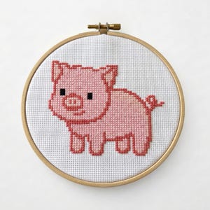 Puede incluir: Un bastidor de bordado de punto de cruz con un diseño de cerdito rosa. El cerdo está representado en un estilo pixelado con un cuerpo rosa claro y un contorno rosa más oscuro. El fondo es blanco y el bastidor es de madera.