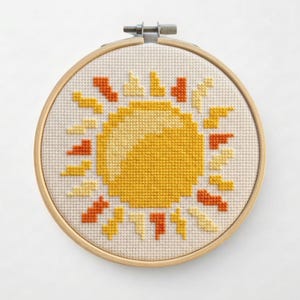May include: A cross-stitch embroidery hoop featuring a yellow sun design with orange and yellow rays. The sun is centered on a cream-colored fabric, framed by a wooden hoop. The design is a cheerful, handmade craft.