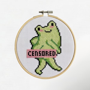 Op de afbeelding: Een borduurring met kruissteek met een lichtgroene kikker met een roze "CENSORED" banner. De kikker heeft zwarte ogen en een kleine glimlach. De borduursel is geplaatst in een houten ring tegen een witte achtergrond.