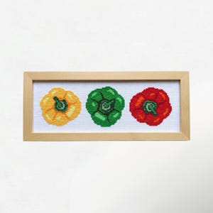 May include: Cross-stitch artwork featuring three bell peppers in yellow, green, and red, displayed in a rectangular wooden frame. The peppers are detailed with stems and are set against a white background.