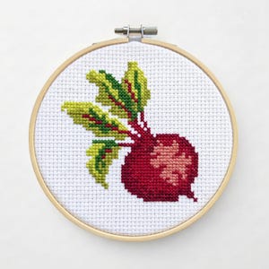 May include: A cross-stitch embroidery of a red beet with green and yellow leaves. The beet is centered on a white fabric, framed by a wooden embroidery hoop. The design features detailed stitching.