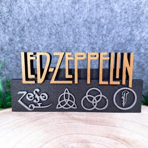 Peut inclure: Panneau en bois avec le nom "LED ZEPPELIN" découpé. Le panneau comprend les symboles du groupe en dessous du nom, sur une base gris foncé. Les lettres sont de couleur marron clair.