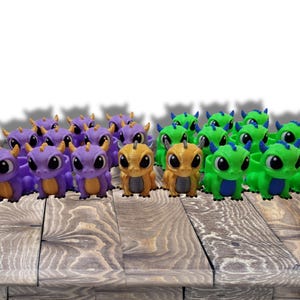 May include: A collection of small, colorful dragon figurines. The dragons are in shades of purple, gold, and green, with large eyes and small horns. They are arranged on a wooden surface.