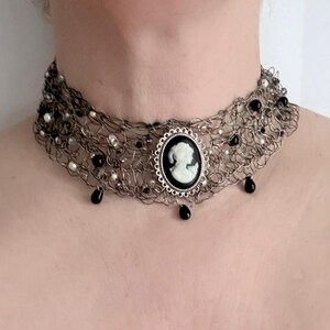 May include: A silver choker necklace with a cameo pendant. The necklace has a woven design with black and white beads and teardrop accents. The cameo pendant features a white profile on a black background.