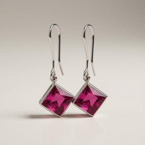 May include: A pair of silver drop earrings featuring square-cut, vibrant magenta-pink gemstones. Each earring is suspended from a simple silver hook wire, showcasing a faceted design that catches the light, creating a clear, polished appearance.