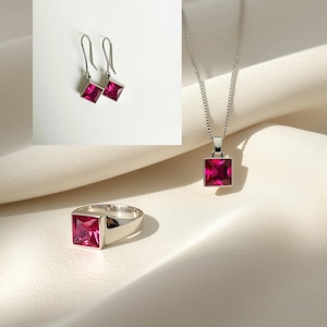 Puede incluir: Un conjunto de joyería compuesto por un anillo de plata, un collar con colgante de plata y un par de pendientes de plata. Cada pieza presenta una piedra preciosa de color magenta de corte cuadrado engastada en un marco de plata pulida.