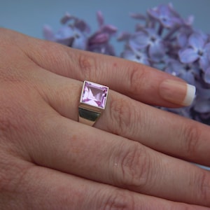 May include: A silver ring featuring a square-cut, light pink gemstone set in a smooth, polished metal band. The stone has a faceted surface that reflects light, and the setting is a simple, clean design that holds the square gem securely in place.