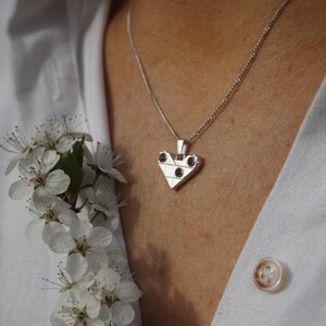 May include: A silver-toned heart-shaped pendant necklace featuring a geometric grid pattern and three small, round, dark red stones set into the metal. The pendant hangs from a thin silver-toned chain against a white collared shirt with a visible button.
