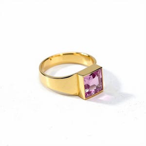 Op de afbeelding: Een gouden ring met een vierkant geslepen, lichtpaarse edelsteen in een gladde, gepolijste band. De rechthoekige zetting houdt de gefacetteerde steen stevig vast, wat zorgt voor een strakke, geometrische uitstraling met een heldere, reflecterende afwerking.
