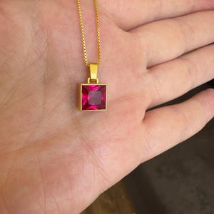 Op de afbeelding: Een goudkleurige boxketting met een vierkante, levendige magenta-roze edelsteen hanger in een gepolijst goudkleurig metalen frame. De hanger hangt aan een fijne, dunne goudkleurige ketting die verbonden is met een eenvoudige, massieve metalen hanger.