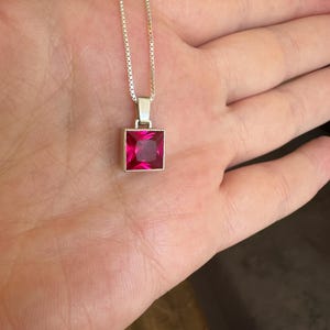 May include: A silver box chain necklace featuring a square-cut, vibrant pink gemstone pendant set in a simple silver frame. The pendant hangs from a delicate, polished silver chain, showcasing a clean, geometric design with a bright, saturated color.