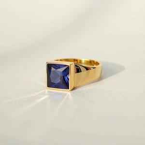May include: A gold-toned signet ring featuring a large, square-cut, deep blue gemstone set in the center. The band is smooth and polished, reflecting light across its surface, while the faceted blue stone creates a sharp, geometric focal point.