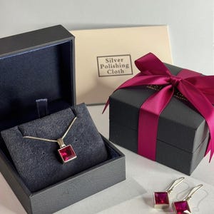 Peut inclure: Un collier en argent avec un pendentif carré rouge et des boucles d'oreilles assorties. Les bijoux sont présentés dans une boîte noire à l'intérieur bleu foncé, avec une boîte cadeau noire fermée par un ruban magenta et une chamoisine.