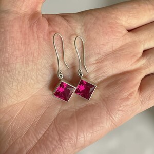 Puede incluir: Un par de pendientes colgantes con gemas de color rosa fucsia vibrante de corte cuadrado, engastadas en monturas de metal plateado pulido. Cada pendiente cuelga de un sencillo gancho de alambre plateado curvado.