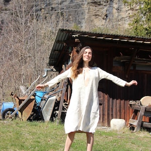 May include: A cream linen dress with a deep V-neck and long sleeves. The dress is knee-length and has a relaxed fit. The person is wearing brown sandals. The background includes a wooden structure, a mountain, and a blue wheelbarrow.