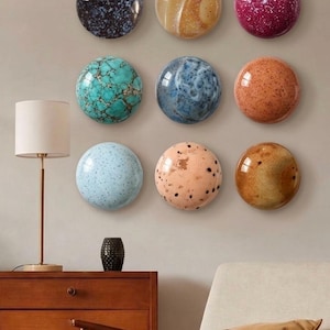 Peut inclure: Une collection de neuf pièces d'art mural circulaires disposées en grille. Chaque pièce présente une finition unique, notamment bleu foncé moucheté, beige marbré, magenta avec des taches blanches, turquoise, marbre bleu, orange, bleu clair, pêche avec des points noirs et marron.