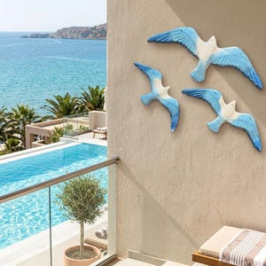 May include: Three decorative seagull wall art pieces with white bodies and blue wings are mounted on a beige wall. The background shows a pool, palm trees, and the ocean. The seagulls are in a flying pose.