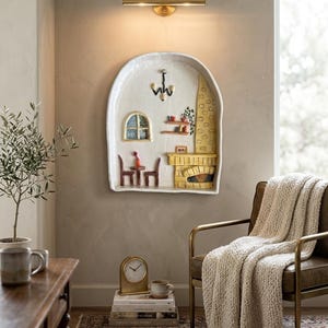 May include: A ceramic wall art piece showing a miniature interior scene with a fireplace, table, and chairs. The piece is white with colourful accents and is mounted on a neutral wall. A brass light fixture is above the ceramic piece.