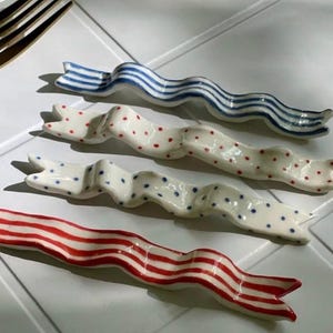 Può includere: Quattro poggiabacchette in ceramica a forma di nastro. Uno è blu a strisce bianche, un altro è bianco a pois rossi, il terzo è bianco a pois blu e l'ultimo è rosso a strisce bianche.