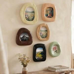 Peut inclure: Un ensemble de six cadres muraux en céramique aux formes organiques irrégulières, déclinés en crème, pêche, marron, beige, noir et vert sauge. Chaque cadre contient une impression différente de paysage ou de fleurs, disposés en groupe.