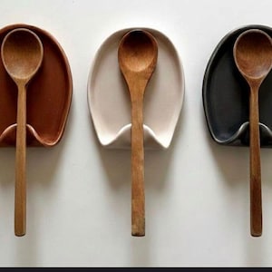 May include: Three ceramic spoon rests in brown, cream, and black, each holding a wooden spoon. The spoon rests have a curved design to cradle the spoon. A simple and functional kitchen accessory.