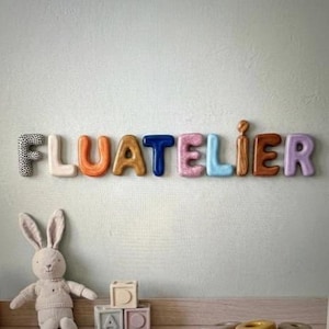 May include: Colorful wooden letters spelling "FLUATELIER" are mounted on a wall. A stuffed bunny and wooden blocks are in the foreground. The letters are various colors and textures, creating a playful and decorative display.
