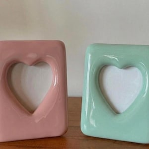Puede incluir: Dos marcos de fotos rectangulares con acabado brillante, uno en rosa pastel y otro en verde menta. Cada marco presenta un recorte central en forma de corazón para mostrar una fotografía, con bordes suaves y redondeados.