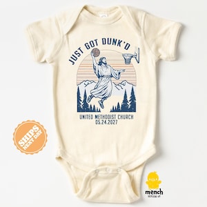 May include: A cream-colored short-sleeve baby bodysuit featuring a blue graphic of a figure dunking a basketball into a hoop. The text reads JUST GOT DUNK'D, UNITED METHODIST CHURCH, and 05.24.2027, set against a background of mountains and pine trees.