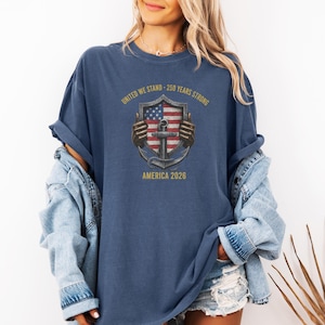 Puede incluir: Camiseta azul marino con un gráfico que presenta una bandera estadounidense, un ancla y el texto "UNITED WE STAND - 250 YEARS STRONG AMERICA 2026". El diseño es en tonos rojos, blancos y azules.