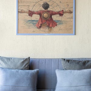 Puede incluir: Obra de arte enmarcada que representa a un personaje de la serie de anime One Piece, con los brazos extendidos, vistiendo una chaqueta roja y un sombrero de paja. El fondo incluye un dise&ntilde;o de mapa y un marco azul. La obra est&aacute; sobre un sof&aacute; azul con cojines grises.