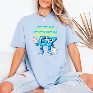Puede incluir: Camiseta azul claro con el texto "SIX SEVEN" en degradado azul y amarillo. Los números 6 y 7 están personificados con brazos, piernas y zapatos. El número 6 hace un signo de la paz. La camiseta es de estilo casual.
