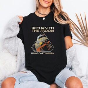 May include: Black t-shirt with the text "RETURN TO THE MOON" in gold and white. Below the text is a graphic of the moon and a triangle with the text "LUNAR FLYBY MISSION". The shirt is a crew neck.