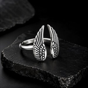 May include: A silver ring featuring an open wing design. The ring is made of polished metal and has intricate feather detailing. The ring is displayed on a dark, textured surface, showcasing its elegant design.