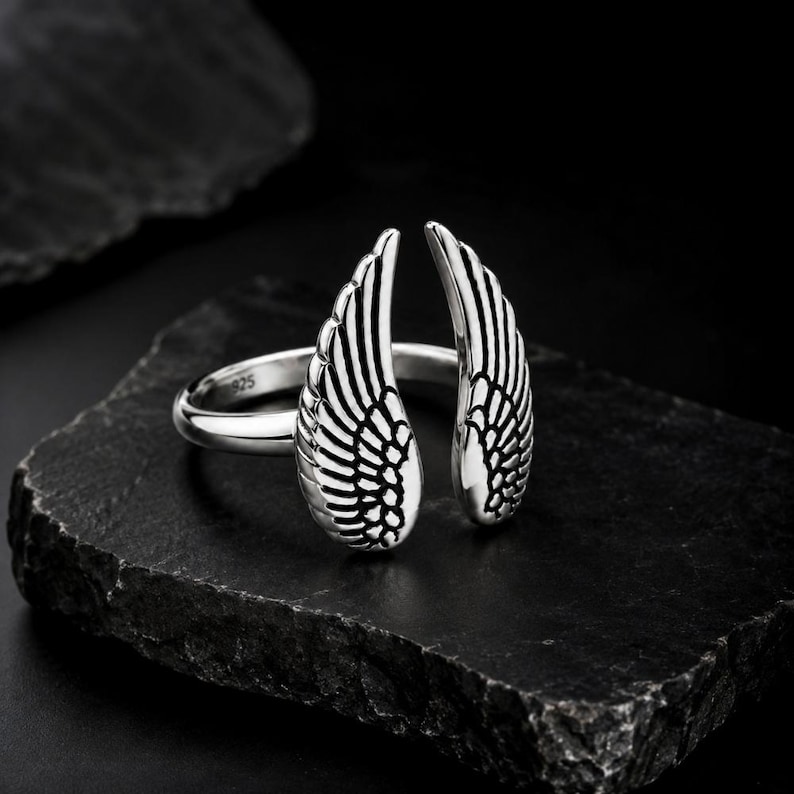 May include: A silver ring featuring an open wing design. The ring is made of polished metal and has intricate feather detailing. The ring is displayed on a dark, textured surface, showcasing its elegant design.