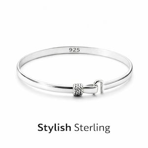 May include: A silver bangle bracelet featuring a hook-and-eye closure mechanism with a twisted rope-textured detail at the center. The interior of the band is clearly stamped with the text 925, indicating sterling silver composition.