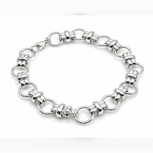 Puede incluir: Una pulsera de cadena en tono plateado con un patrón repetitivo de eslabones circulares conectados por segmentos de doble barra más pequeños. La pulsera tiene un acabado metálico pulido e incluye un pequeño cierre de mosquetón.