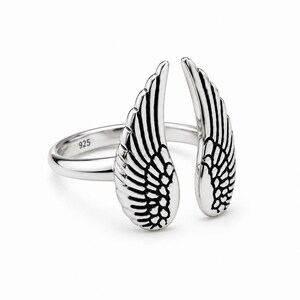 May include: A silver ring featuring an open wing design. The ring has two detailed wings, each with intricate feather detailing. The band is smooth and has the number "925" stamped on it. The ring is set against a white background.