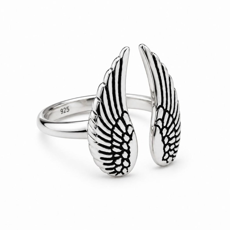 May include: A silver ring featuring an open wing design. The ring has two detailed wings, each with intricate feather detailing. The band is smooth and has the number "925" stamped on it. The ring is set against a white background.
