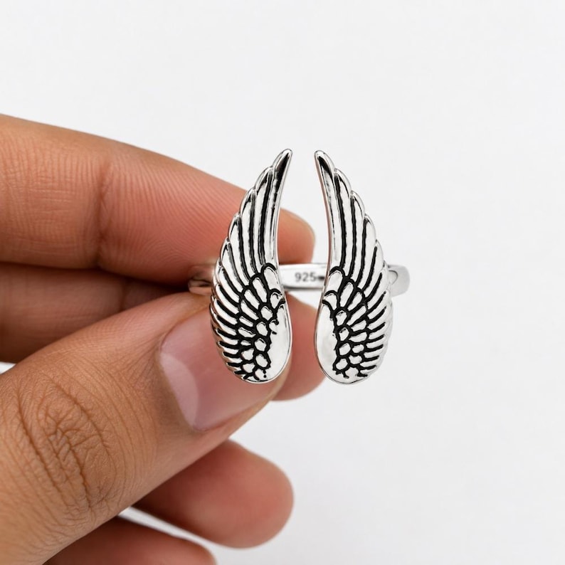 May include: A silver ring featuring an open design with two detailed wing shapes. The ring is marked with "925" and is held by a hand against a neutral background. The wings have intricate feather detailing.