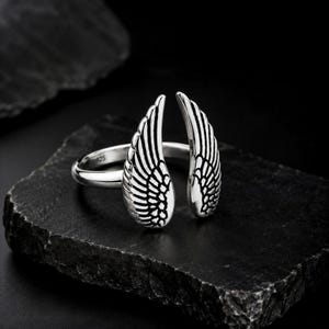 May include: A silver ring featuring an open wing design. The ring is made of polished metal and has intricate feather detailing. The ring is displayed on a dark, textured surface, highlighting the contrast between the shiny metal and the dark background.
