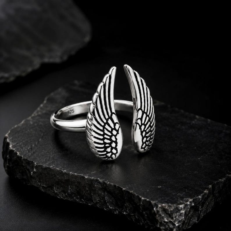 May include: A silver ring featuring an open wing design. The ring is made of polished metal and has intricate feather detailing. The ring is displayed on a dark, textured surface, highlighting the contrast between the shiny metal and the dark background.