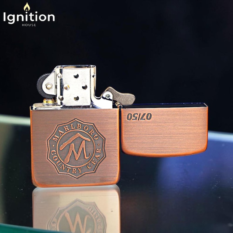 May include: A copper-colored metal lighter with the text "MARLBORO COUNTRY GEAR" and a stylized "M" logo. The lighter is open, revealing the interior mechanism and the text "OSIZO". The top of the lighter is silver.