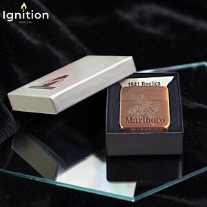 May include: A copper-colored 1941 Replica Marlboro lighter with a cowboy design, presented in a black box. The lighter is displayed next to its silver box, with the "Ignition House" logo in the top left corner.