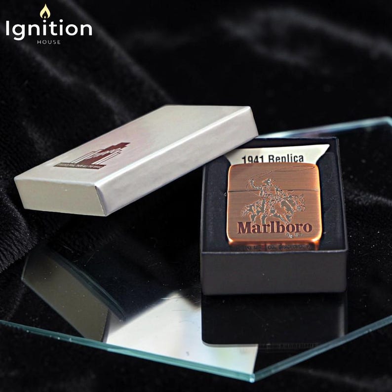 May include: A copper-colored 1941 Replica Marlboro lighter with a cowboy design, presented in a black box. The lighter is displayed next to its silver box, with the "Ignition House" logo in the top left corner.