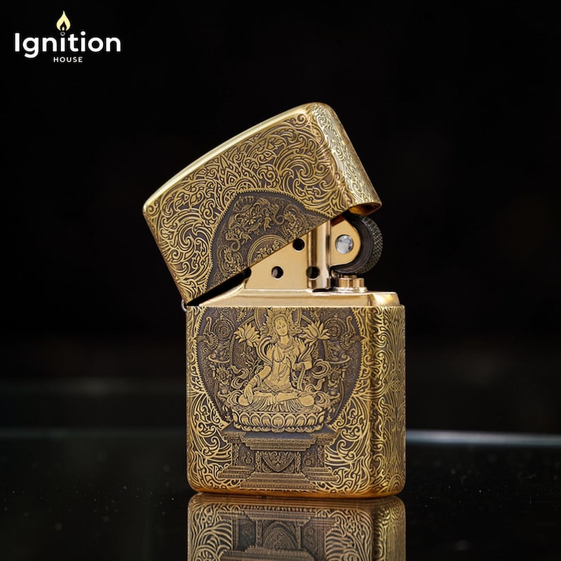 May include: Gold-colored metal lighter with an open lid. The lighter features intricate engraved patterns and a central image of a seated figure. The text "Ignition House" is in the upper left corner.