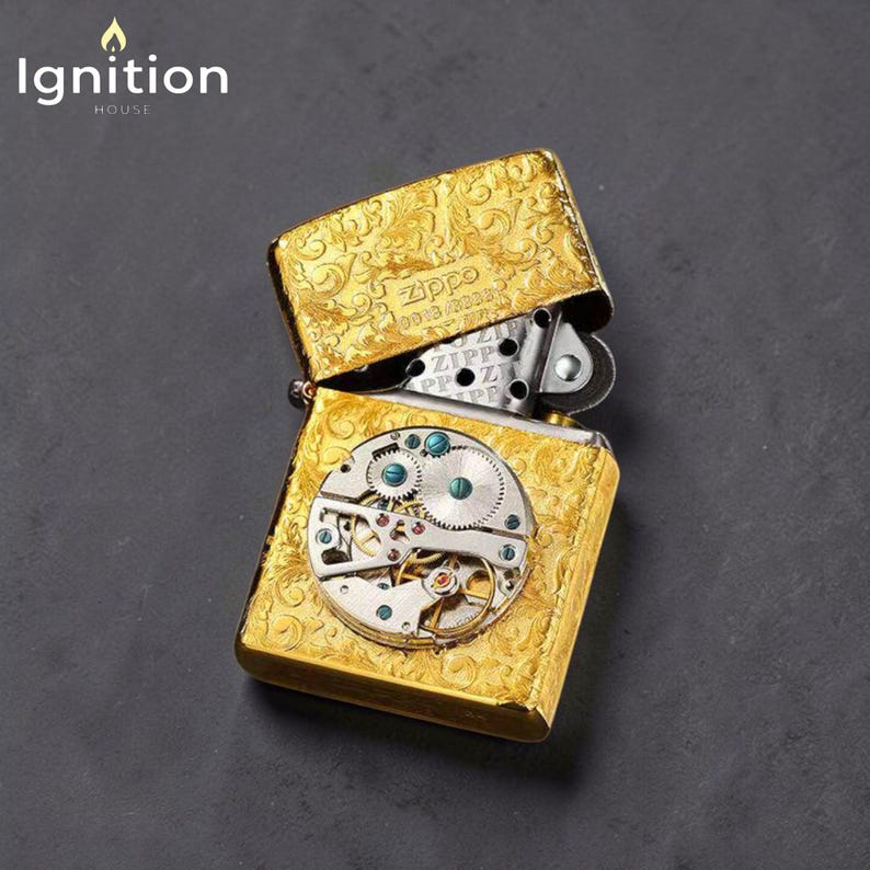 Puede incluir: Encendedor Zippo dorado con tapa abierta, que revela intrincados detalles de relojer&iacute;a. El encendedor presenta un patr&oacute;n floral grabado. Los textos "Zippo" e "Ignition House" son visibles.