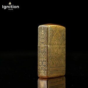 May include: A gold-colored lighter with intricate engraved patterns. The lighter has a rectangular shape with rounded edges and a hinged lid. The design includes floral and geometric motifs. The brand name "Ignition House" is in the top left corner.
